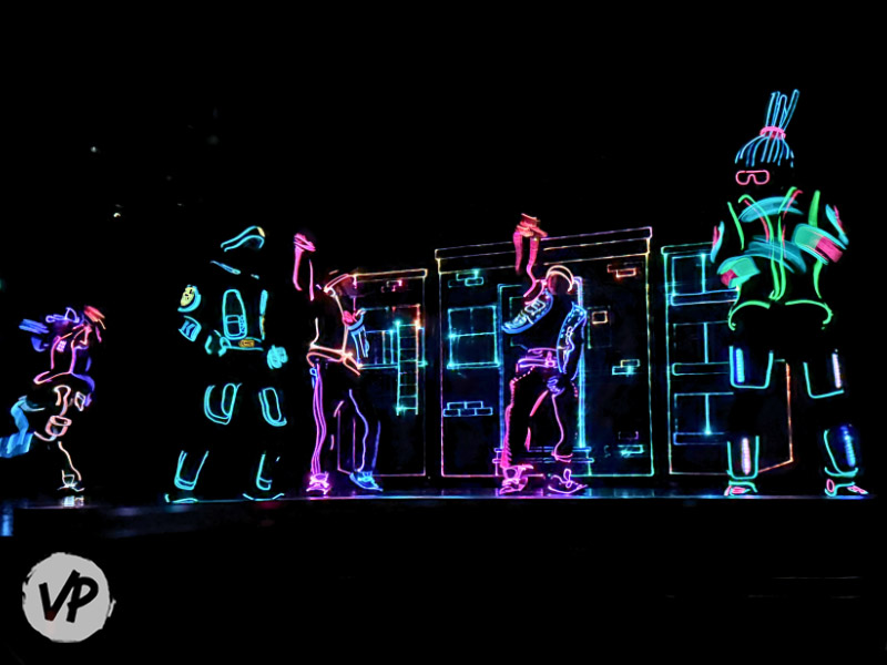 The performers dance in the dark to a song, wearing electric glow-in-the-dark suits
